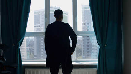 Rear view of a dressed man standing indoors in a living room by the window. Media. Silhouette of a...