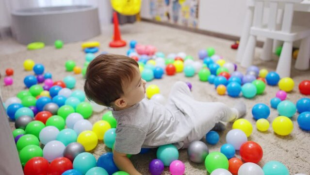 Cute kid sits on the floor among the scattered balls. Toddler lies down on the balls and tumbles by them.
