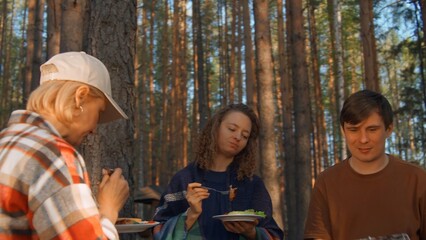 Fototapeta premium Friends eat barbecue from grill in woods. Stock footage. Friends relax in nature at barbecue and eat. Friends eat cooked meat on grill on sunny summer day