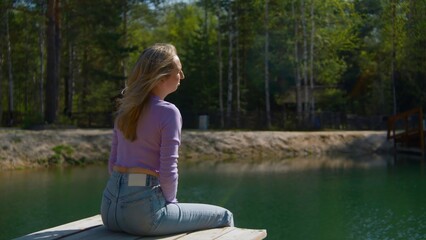 Young woman sits on pier and throws stone. Stock footage. Beautiful young woman sits on edge of pier by lake in summer. Woman on lake pier on sunny summer day