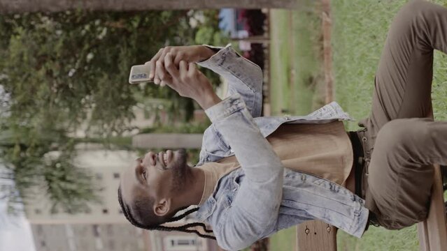 Young black man, sitting on the bench in the square, smiling, taking a selfie with his smartphone. Cinematic 4k. Vertical video.