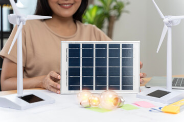 Cropped view of woman holding light bulb on white table with model of wind turbine, solar cell and...