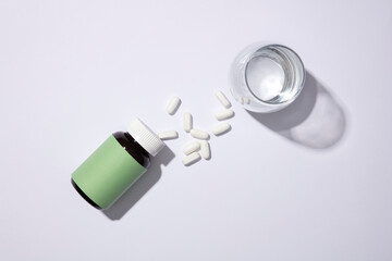 Top view of white tablets, an unbranded medicine bottle and a glass of water on white background. Medical theme for medicine advertising. Minimalist background.