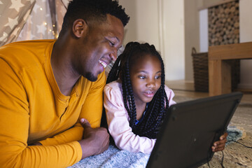 Happy african american father and daughter lying by teepee with computer at home, copy space