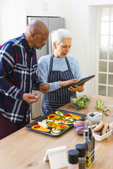 Diverse senior couple preparing healthy meal with vegetables using tablet in kitchen