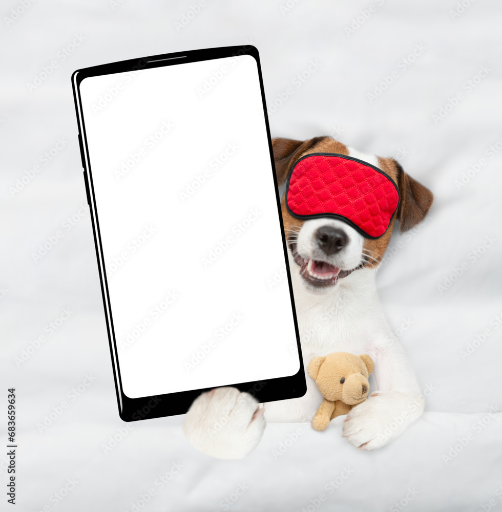 Poster jack russell terrier puppy wearing sleeping mask lying on a bed at home and showing big smartphone w