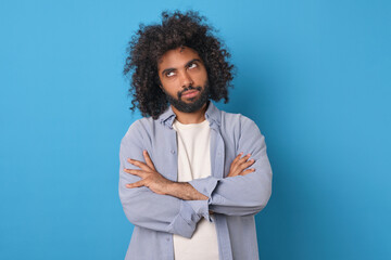 Young doubtful funny Arabian man rolls eyes and looks up with offense crossing arms in front of chest after insult or inappropriate remark from boss posing on isolated blue background. - obrazy, fototapety, plakaty
