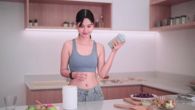 Asian sportswoman in sportswear having a protein shake to manage weight in her home kitchen. She focuses on maintaining good health, consuming nutritious food, and keeping a positive body image.