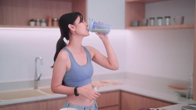 Asian sportswoman in sportswear having a protein shake to manage weight in her home kitchen. She focuses on maintaining good health, consuming nutritious food, and keeping a positive body image.