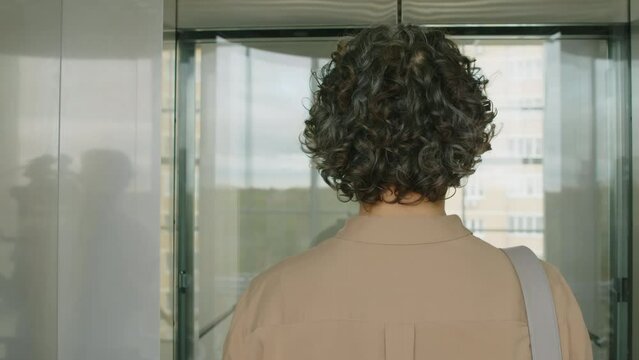 Back tracking shot of unrecognizable businesswoman with curly hair and in officewear getting off panoramic elevator and going towards work room inside modern office buildingBack tracking shot of unrec