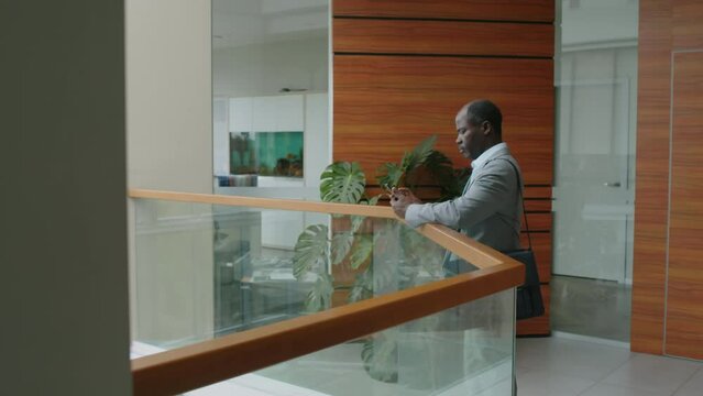 Side slowmo of African American businessman in grey suit with briefcase coming to railing in office atrium and making phone callSide slowmo of African American businessman in grey suit with briefcase 