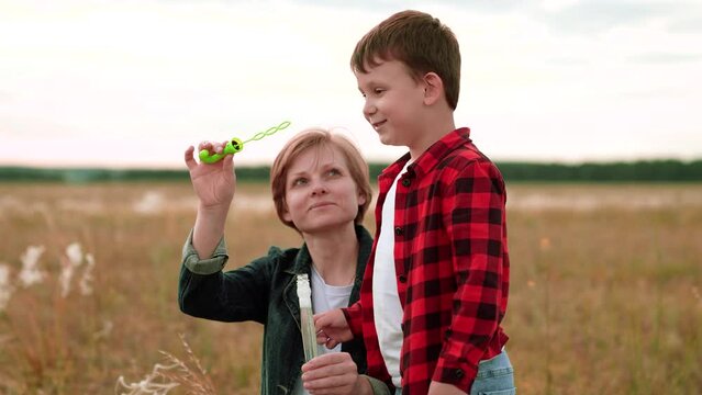 Boy in checkered shirt blows bubbles with mother help on freshly mowed field