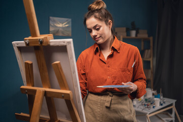 Young beautiful painter woman standing in modern workshop painting picture on canvas with paints, enjoy creative hobby. Education, and art-class