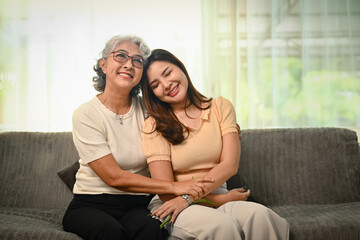 Obraz premium Smiling grey haired elderly woman embracing her grownup daughter sitting on sofa at home