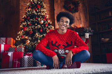 Photo of cheerful positive guy wear red ugly xmas sweater sitting under christmas tree indoors apartment room