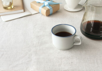 Coffee making scenery including a table with a gray tablecloth with natural taste, presents, white mugs and coffee server.