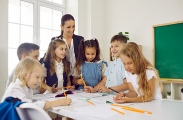 Group of elementary students learning new things in class. Happy children and teacher gather around table in classroom, write in notebooks, draw pictures, and discuss stories. Back to school concept