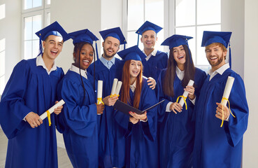 Diverse group of a young joyful people wearing blue graduation gowns indoors looking cheerful at camera with diplomas in hands. Happy graduate students portrait. Education concept.