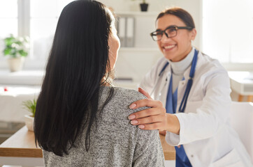 Obraz premium Portrait of smiling friendly doctor support female patient putting hand on her shoulder. Physician wearing stethoscope giving consultation a woman sitting back during medical examination in clinic.