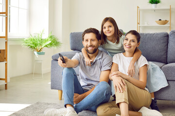 Happy nuclear family watching TV at home. Mother, father and child daughter sitting on floor by sofa in living room interior, looking away, watching television show together and smiling