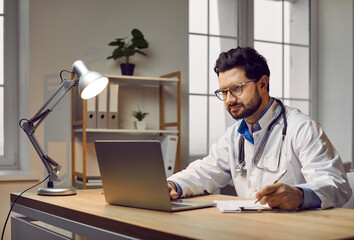 Happy man doctor in white medical coat with stethoscope sitting at desk with lamp in office...