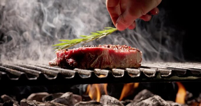 Smell of juicy meat sizzling over hot coals is synonymous with summer. Summer parties come alive with smoky aroma of delicious grilled steak. Hand puts sprig of fragrant rosemary on steak.