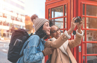 Fototapeta na wymiar two friends, girlfriend and women using a mobile phone, camera and taking a selfie against a red phonebox in a city in England. Travel Lifestyle concept