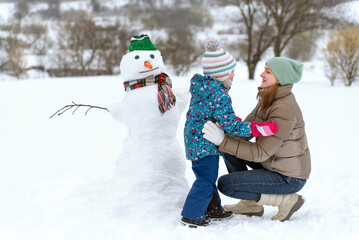 Young mother and child next to large snowman have fun. Winter weekend with the family outside the city. - obrazy, fototapety, plakaty