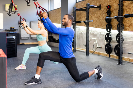 A couple of different ethnicities perform some exercises with special tapes inside a gym. The young people work their legs by doing one-foot squats with suspension straps.