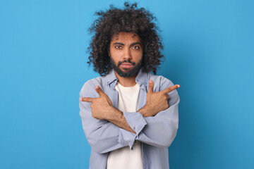 Young handsome casual Arabian man points with index fingers of both hands in different directions recommending that you pay attention to advertised product stands posing in blue studio. - obrazy, fototapety, plakaty