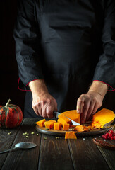 A male chef uses a knife to cut an orange pumpkin into slices for baking on a wooden table. Autumn...