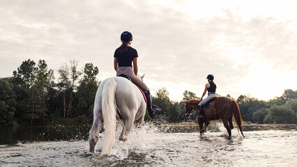 Two rider girls crossing the calm river water riding their beautiful horses on the cloudy summer...