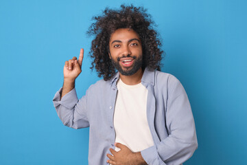Young cheerful Arabian man student experiences stomach pain from laughing and points finger up inviting him to attend stand-up show with popular comedians stands posing in blue studio.