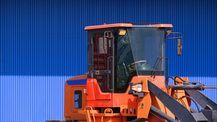 Cab control room of orange hydraulic excavator in front of blue warehouse building wall in construction site, close up with copy space