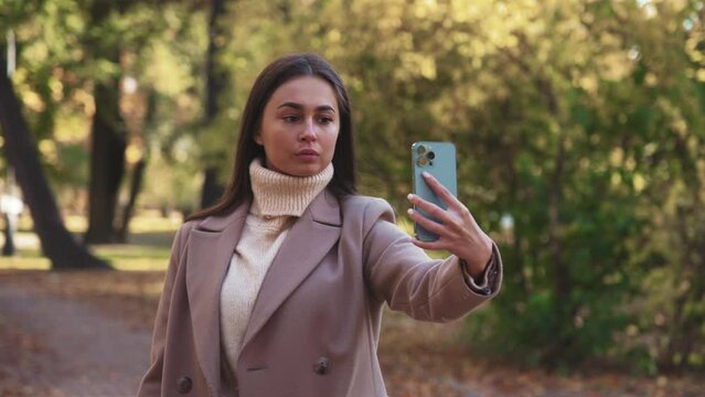 Young beauty woman walks in coat in autumn park, holds phone in hand and takes picture of herself on front camera and takes selfie or talks and communicates on video call. Leisure in nature landscape.