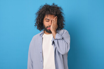 Young tired ethnic Arabian man covers face with palm feeling loss of strength and lack of opportunity for further action due to problems in personal life stands posing in blue studio.