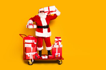 Full size portrait of elderly santa claus stand market pushcart hold new year giftbox empty space isolated on yellow color background
