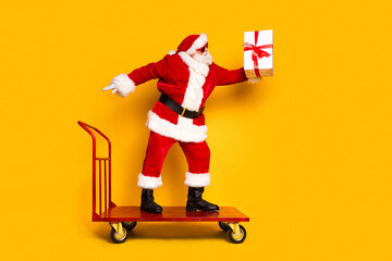 Full length profile portrait of elderly santa supermarket pushcart give christmas giftbox empty space isolated on yellow color background