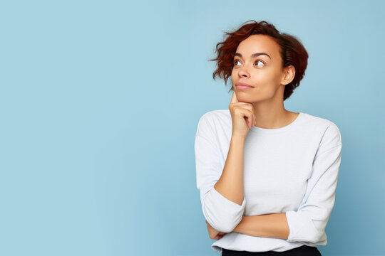 Jeune femme en train de penser en regardant sur le côté vers le haut, espace libre pour du texte ou mise en scène, tenue claire fond uni bleu pâle