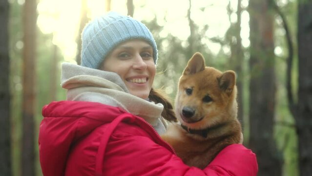 Portrait of a young woman in a red jacket and a blue hat and a Shiba Inu puppy in a close-up in a forest park. They look at the camera, look at each other. dog is nuzzling its owner. Female laughter