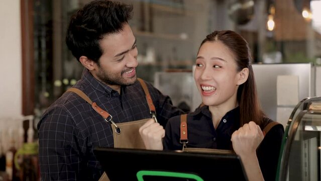 Two cheerful baristas running coffee shop standing behind bar counter and professional machine exciting first online order using modern technology POS machine,  Asian and Indian couple business owner