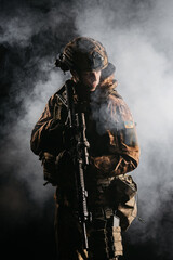 Fototapeta premium An army soldier in a military camouflage uniform, a helmet and a mask, holds a rifle and aims with a red dot sight, standing in an attack position, in the smoke on a black background.