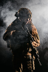 An army soldier in a military camouflage uniform, a helmet and a mask, holds a rifle and aims with a red dot sight, standing in an attack position, in the smoke on a black background.