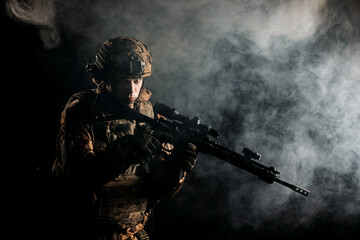 An army soldier in a military camouflage uniform, a helmet and a mask, holds a rifle and aims with...