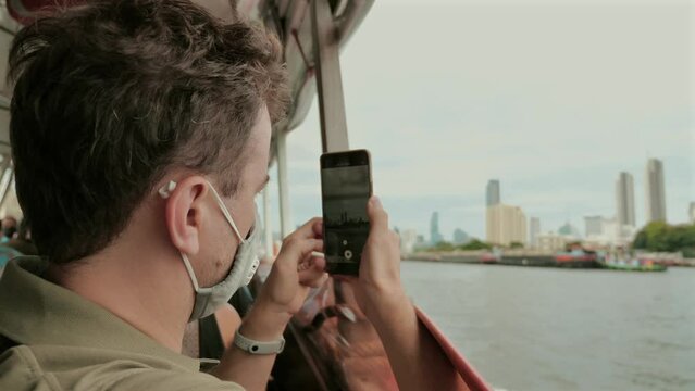 Man wearing face mask taking photo with smartphone on river ferry. Urban exploration and technology.