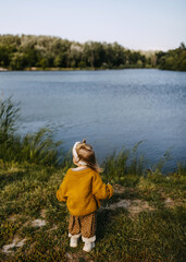 Naklejka na ściany i meble Little girl alone standing by the lake on a sunny day, watching it.