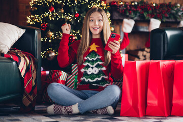 Photo teen shopaholic girl sitting on carpet raised fist up browsing smartphone finally found desirable xmas postcard gift isolated indoors