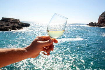 A man's hand holds a glass of champagne against the backdrop of the sea and mountains. Vacation concept - 683808014