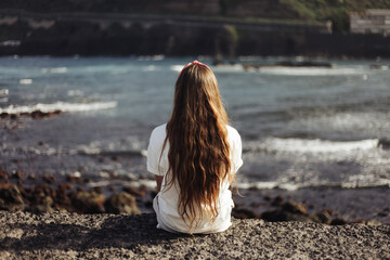 Fototapeta na wymiar Woman with long hair watching waves. Sitting on a ocean shore. Tenerife volcanic beach. Canary island sunset. Girl sit edge of a cliff. Contemplation and meditation background. Young girl outdoor.