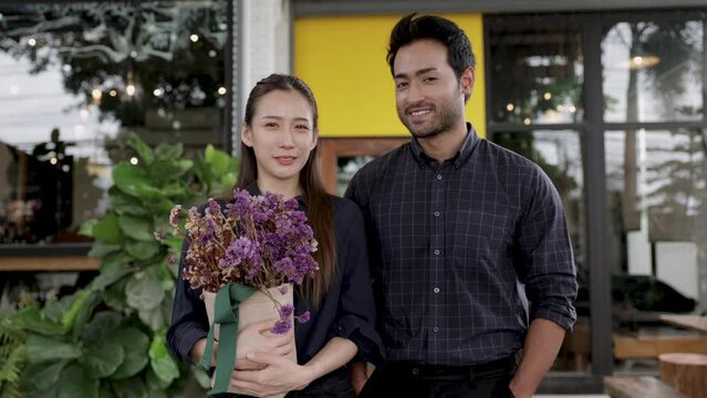 Two cheerful baristas multiethnicity man and woman running coffee shop business entrepreneurship standing in front of cafeteria with flowers smiling, Asian and Indian couple small business strat up